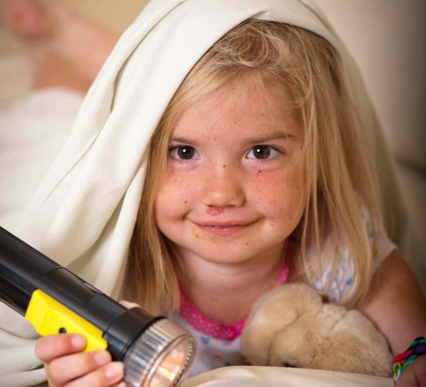 Makenna, a young girl with freckles, smiling under a blanket and holding a flashlight