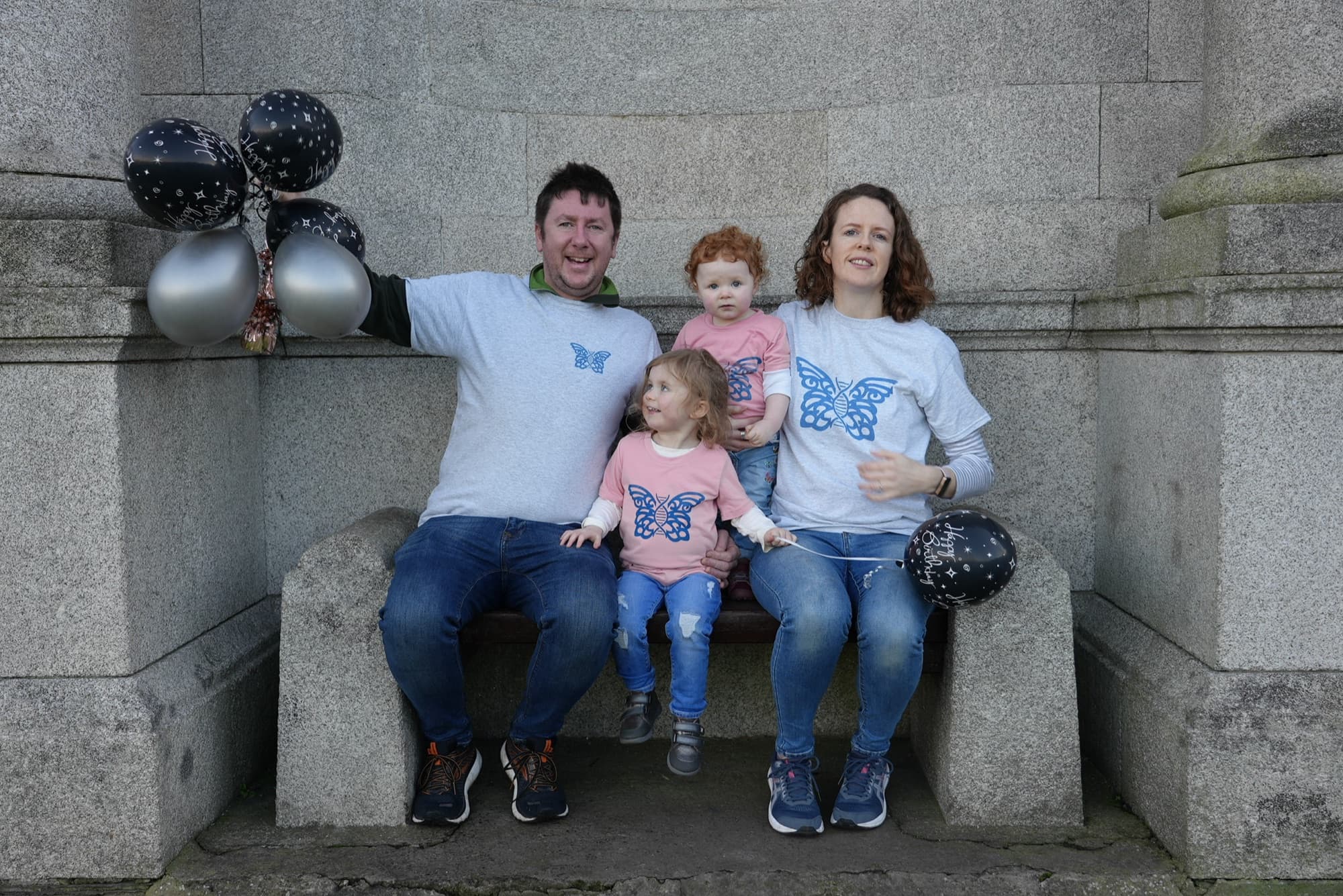 A family of four in matching Cure Blau butterfly T-shirts, holding balloons on a stone bench