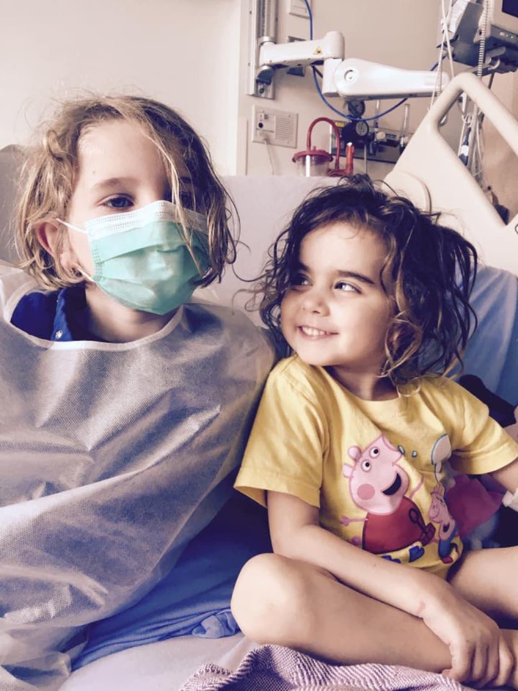 Two young children sitting together on a hospital bed, the older one wearing a mask and gown