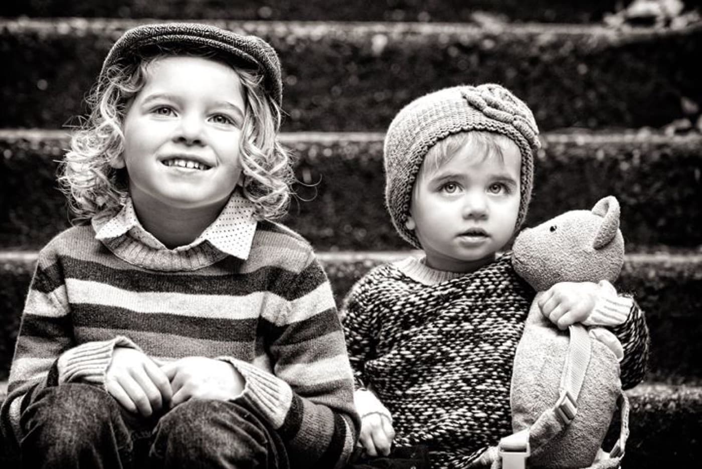 Two young children in knit hats sitting on stone steps, the younger one holding a teddy bear