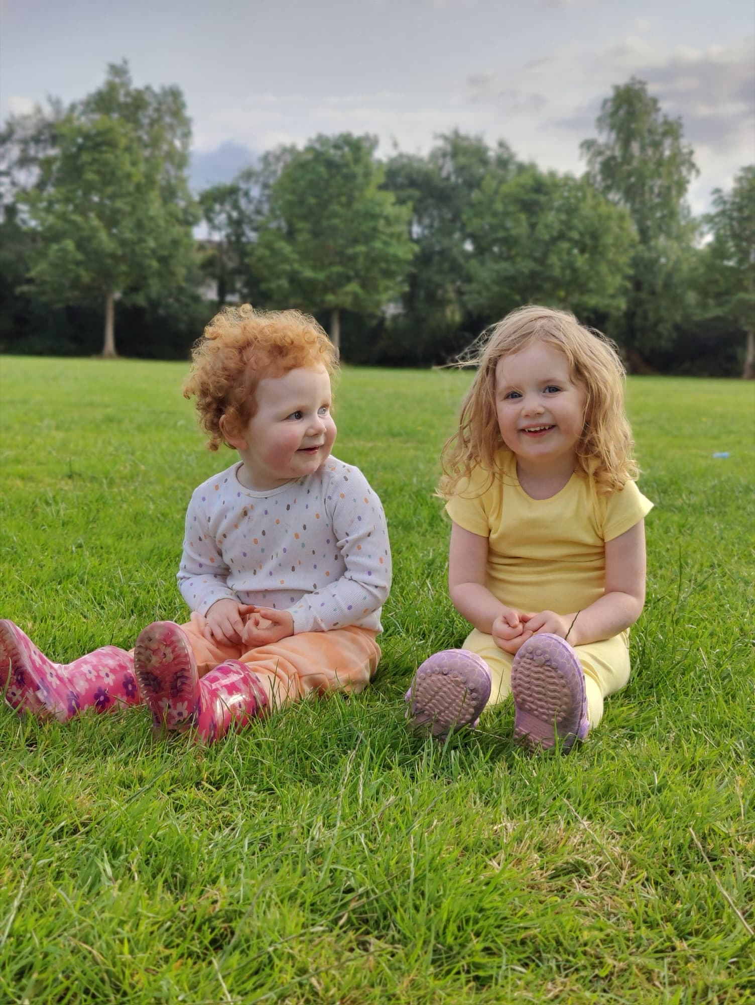 Two small sisters, one with red curls and one with blond hair, sitting on the grass in a park
